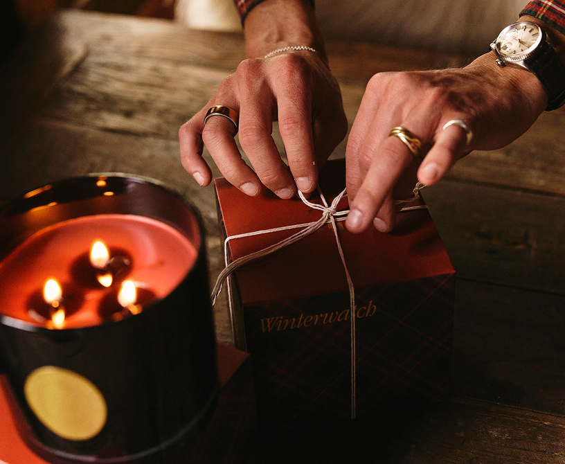 Person tying a string around a gift box with a candle in the foreground on a wooden surface.